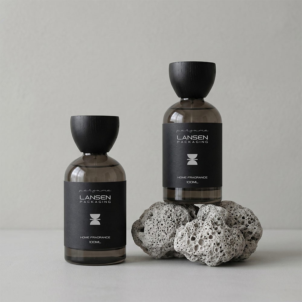 two grey glass perfume bottles with black flared wooden caps and dark wrap labels standing on a light stone surface
