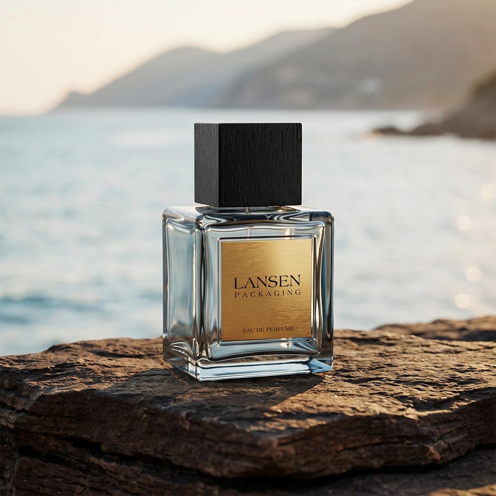 100ml square glass perfume bottle with a brushed gold metal label and a black wooden square cap, standing on rock beside the sea