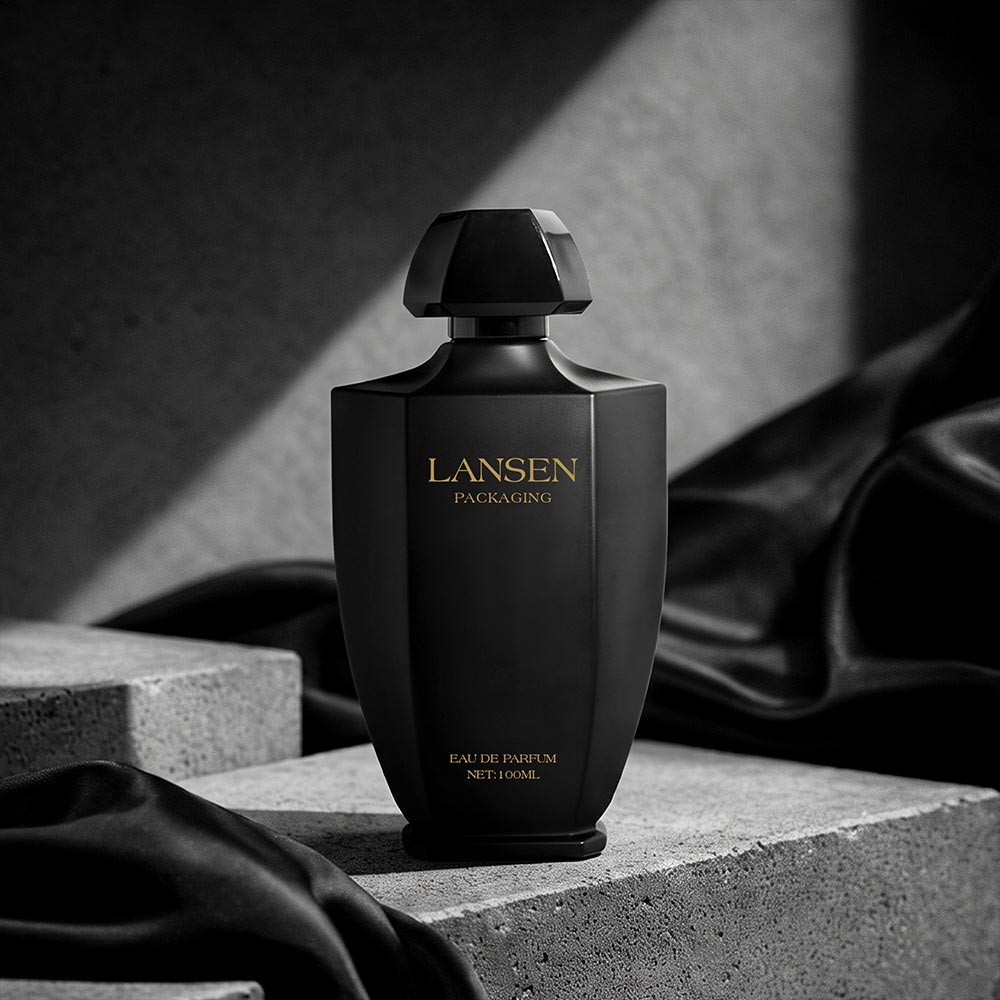 100ml tapered perfume bottle with a matt black faceted body and a gold logo, standing on concrete blocks